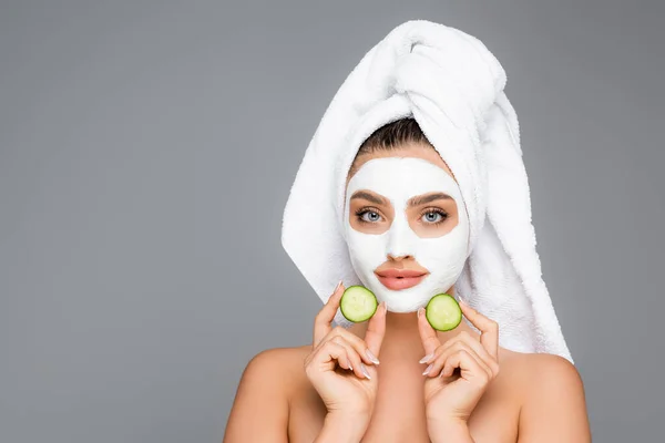 Woman with towel on head and clay mask on face holding cucumber isolated on grey — Stock Photo