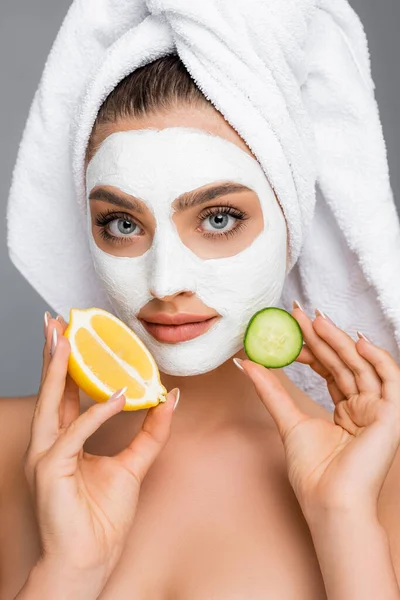 Woman with towel on head and clay mask on face holding lemon and cucumber isolated on grey — Stock Photo