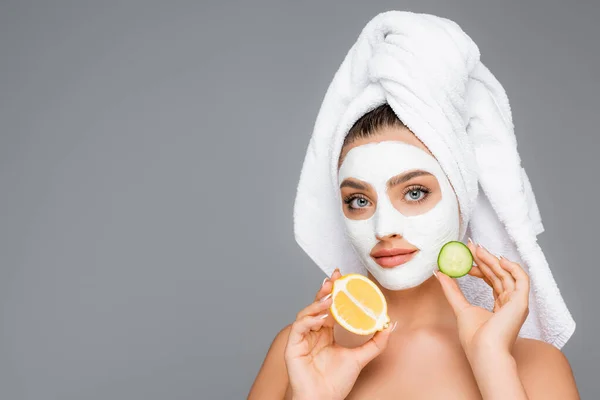 Woman with towel on head and clay mask on face holding lemon and cucumber isolated on grey — Stock Photo