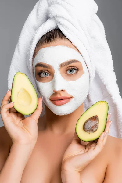 Frau mit Handtuch auf dem Kopf und Tonmaske im Gesicht, die Avocadohälften isoliert auf grau hält — Stockfoto