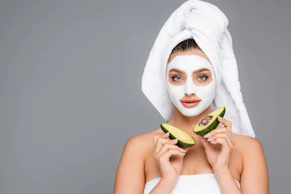 Frau mit Handtuch auf dem Kopf und Tonmaske im Gesicht, die Avocadohälften isoliert auf grau hält — Stockfoto