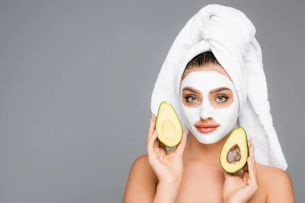 Frau mit Handtuch auf dem Kopf und Tonmaske im Gesicht, die Avocadohälften isoliert auf grau hält — Stockfoto