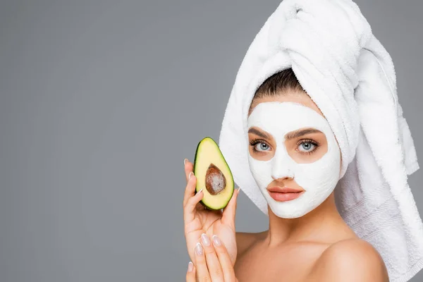 Donna con asciugamano sulla testa e maschera di argilla sul viso tenuta avocado isolato su grigio — Foto stock