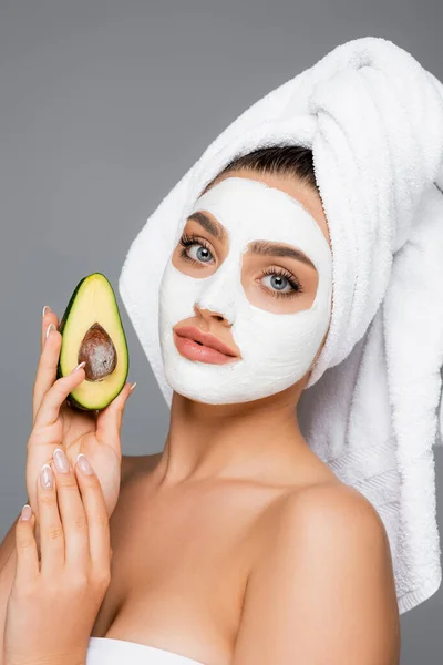 Frau mit Handtuch auf dem Kopf und Tonmaske auf dem Gesicht, die Avocado isoliert auf grau hält — Stockfoto