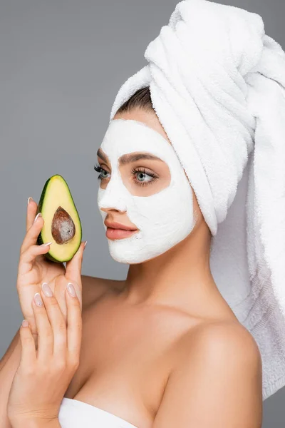 Donna con asciugamano sulla testa e maschera di argilla sul viso tenuta avocado isolato su grigio — Foto stock