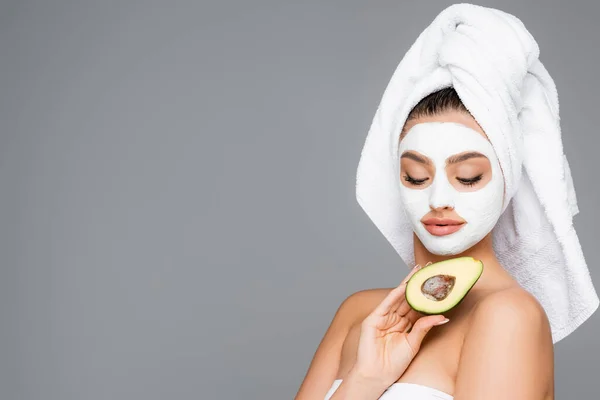 Женщина с полотенцем на голове и глиняная маска на лице держа авокадо изолированы на сером — стоковое фото
