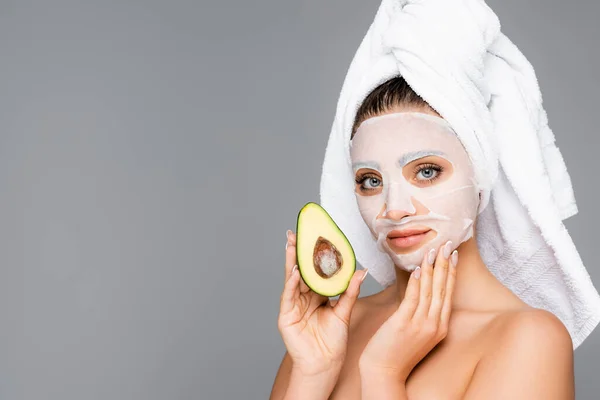 Frau mit Handtuch auf dem Kopf und Maske Laken im Gesicht hält Avocado isoliert auf grau — Stockfoto