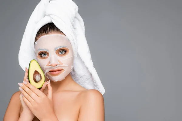 Donna con asciugamano sulla testa e lenzuolo maschera sul viso tenuta avocado isolato su grigio — Foto stock