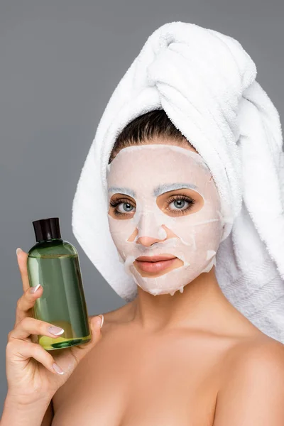 Mujer con toalla en la cabeza y mascarilla en la cara sosteniendo botella aislado en gris - foto de stock