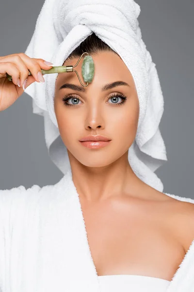 Femme avec serviette sur la tête à l'aide d'un rouleau de jade sur le visage isolé sur gris — Photo de stock