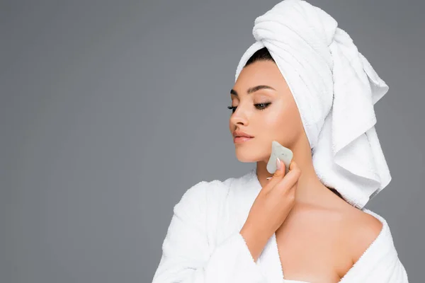Donna con asciugamano sulla testa utilizzando gua sha sul viso isolato sul grigio — Foto stock