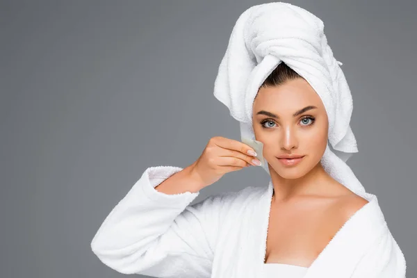 Mujer con toalla en la cabeza usando gua sha en la cara aislada en gris - foto de stock