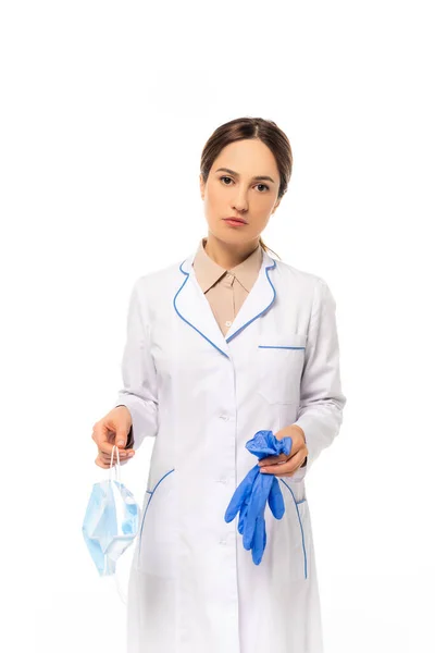 Médico sosteniendo máscara médica y guantes de látex mientras mira la cámara aislada en blanco - foto de stock