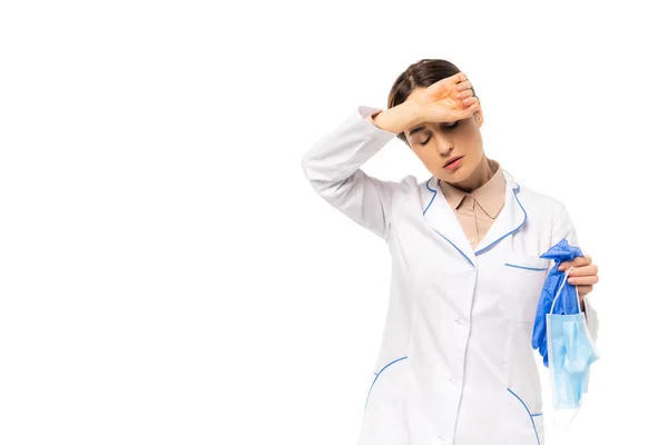 Médico cansado con abrigo blanco sosteniendo guantes de látex y máscara médica aislado en blanco - foto de stock