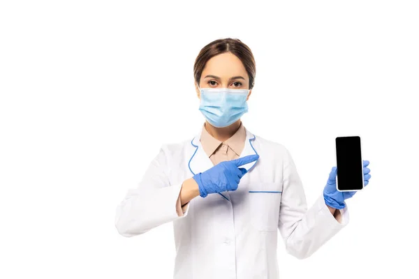 Médico en máscara médica y guantes de látex apuntando con el dedo al teléfono inteligente aislado en blanco - foto de stock