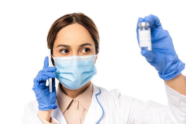 Enfoque selectivo del médico en máscara médica y guantes de látex hablando en el teléfono inteligente mientras mantiene aislada la vacuna en blanco - foto de stock