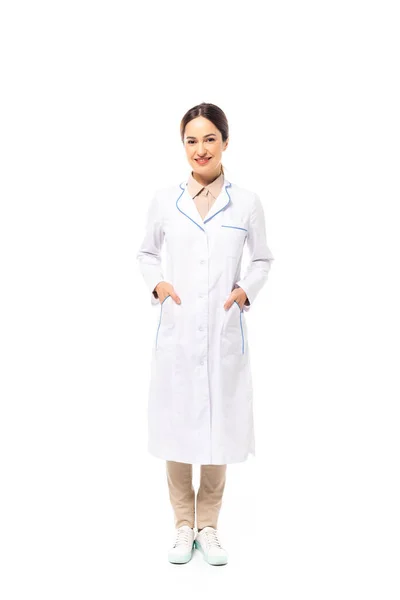 Médico con las manos en bolsillos de abrigo blanco sonriendo a la cámara sobre fondo blanco - foto de stock
