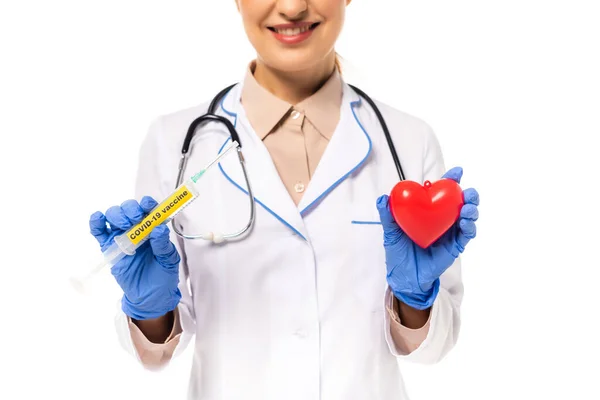 Ausgeschnittene Ansicht eines lächelnden Arztes in Latex-Handschuhen, der eine Spritze mit Covid-19-Impfstoff und Herz auf weiß isoliert hält — Stockfoto
