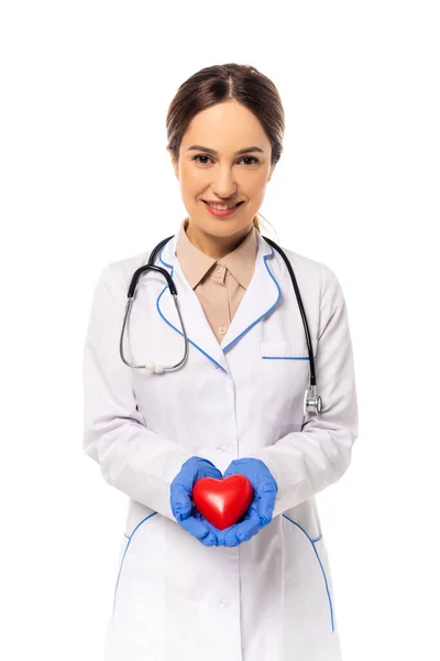 Médico sonriente con guantes de látex que sostiene el corazón rojo aislado en blanco - foto de stock