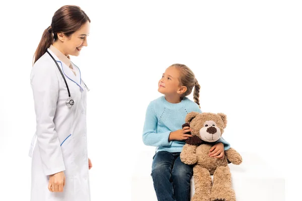 Doctor smiling at girl with teddy bear on table isolated on white — Stock Photo