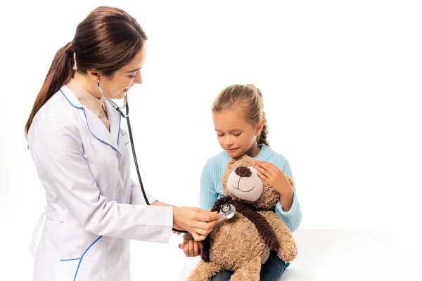 Sonriente médico examinando con estetoscopio juguete suave cerca de chica aislada en blanco - foto de stock