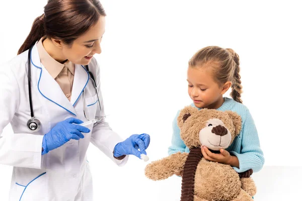 Médico en guantes de látex sosteniendo algodón y jeringa cerca de chica con peluche latido aislado en blanco - foto de stock