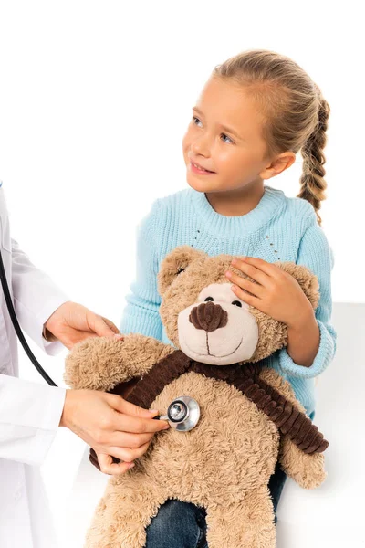 Niño sonriente sosteniendo juguete suave cerca del médico con estetoscopio aislado en blanco - foto de stock