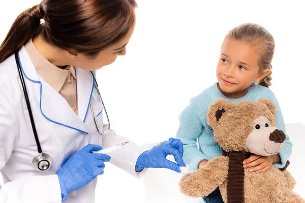 Enfoque selectivo del pediatra en guantes de látex que sostienen algodón y jeringa cerca del niño con juguete suave aislado en blanco - foto de stock