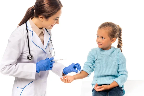 Pediatra sonriente sosteniendo algodón y jeringa cerca de un niño aislado en blanco - foto de stock