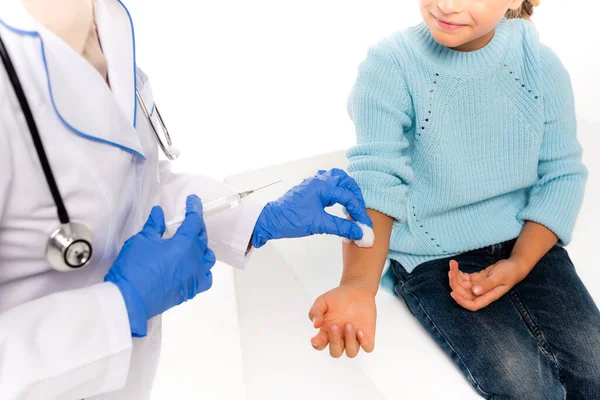 Vista ritagliata del pediatra in guanti di lattice che tengono il cotone e la siringa vicino alla mano del bambino isolato su bianco — Foto stock