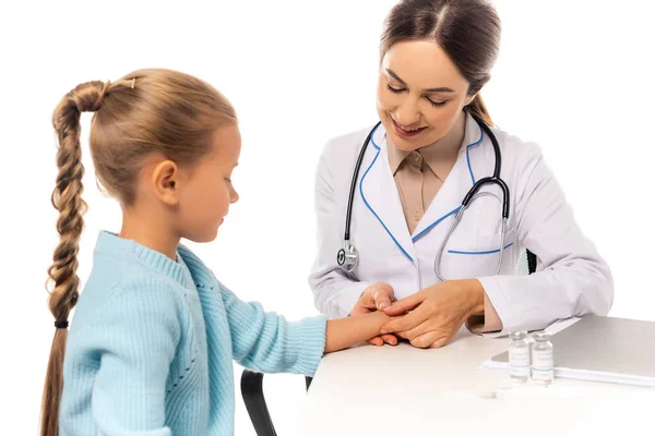 Selektiver Fokus eines lächelnden Arztes, der die Hand eines Kindes in der Nähe von Impfstoff und Papiermappe auf einem Tisch berührt, isoliert auf weiß — Stockfoto