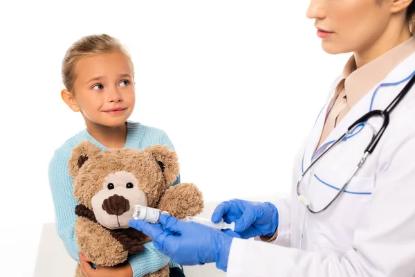 Selektiver Fokus des lächelnden Mädchens mit Teddybär in der Nähe des Arztes mit Spritze und Impfstoff isoliert auf weiß — Stockfoto