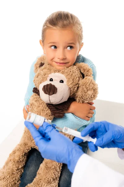 Вибірковий фокус усміхненої дівчини, що тримає м'яку іграшку біля педіатра з вакциною та шприцом ізольовано на білому — стокове фото