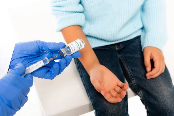 Vista recortada del médico recogiendo la vacuna contra el coronavirus en jeringa cerca de un niño aislado en blanco - foto de stock