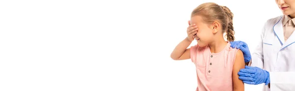 Foto panorámica de niño asustado cubriendo los ojos cerca del médico con guantes de látex aislados en blanco - foto de stock