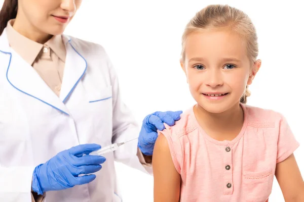 Enfoque selectivo de la niña mirando a la cámara cerca del médico con jeringa aislada en blanco - foto de stock