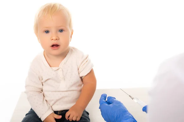 Selektiver Fokus des Kleinkindes, das mit weißer Spritze in die Kamera in der Nähe des Arztes blickt — Stockfoto