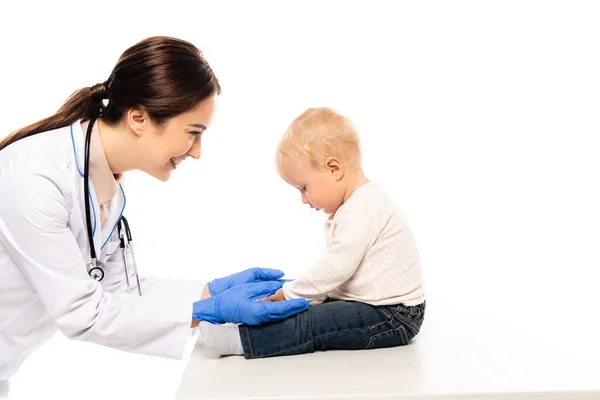 Sorridente pediatra toccare bambino ragazzo sul tavolo isolato su bianco — Foto stock