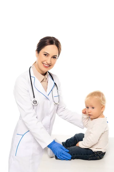 Smiling pediatrician looking at camera near toddler on table isolated on white — Stock Photo
