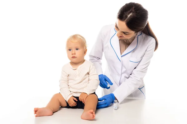 Doctor holding syringe with vaccine near toddler boy on table isolated on white — Stock Photo