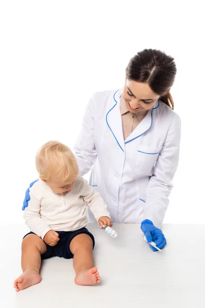Усміхнений лікар тримає шприц біля малюка з баночкою вакцини на столі ізольовано на білому — стокове фото