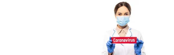 Panoramic shot of doctor in medical mask and latex gloves holding card with coronavirus lettering isolated on white — Stock Photo