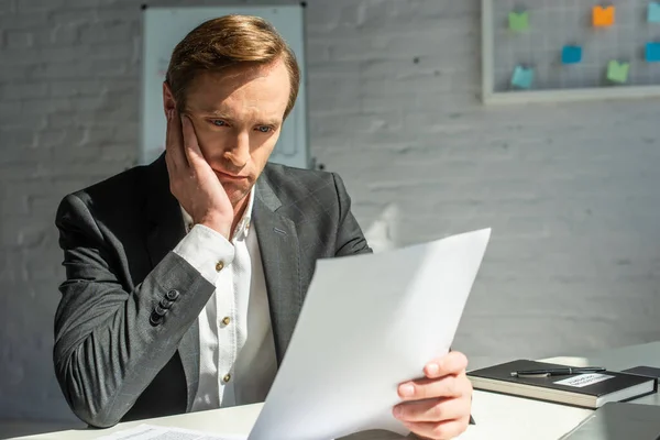 Trauriger Geschäftsmann schaut auf Geschäftsdokument, während er am Arbeitsplatz vor verschwommenem Hintergrund sitzt — Stockfoto