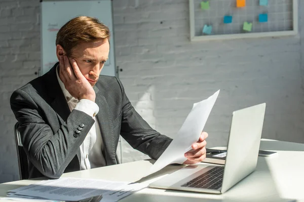 Sad businessman looking at document while sitting at workplace on blurred background — Stock Photo