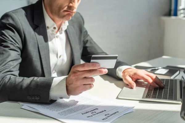 Vista recortada del triste hombre de negocios con tarjeta de crédito, escribiendo en el ordenador portátil, mientras está sentado en el lugar de trabajo en un fondo borroso - foto de stock