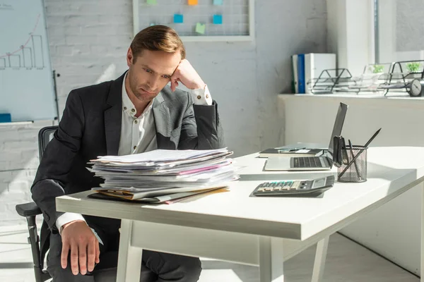 Разочарованный бизнесмен, сидящий на офисном стуле, опираясь на стол с кучей документов — стоковое фото