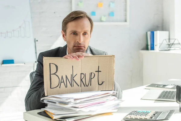 Sad businessman leaning on cardboard with bankrupt lettering, while sitting at table with pile of papers on blurred background — Stock Photo