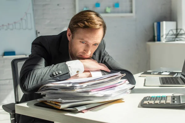 Homme d'affaires déçu appuyé sur une pile de papiers, assis sur le lieu de travail sur un fond flou — Photo de stock