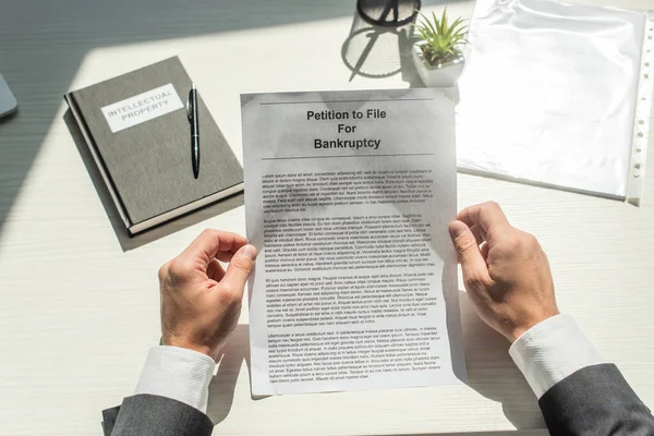 Vista cortada de empresário segurando petição de falência perto notebook em fundo branco texturizado — Fotografia de Stock