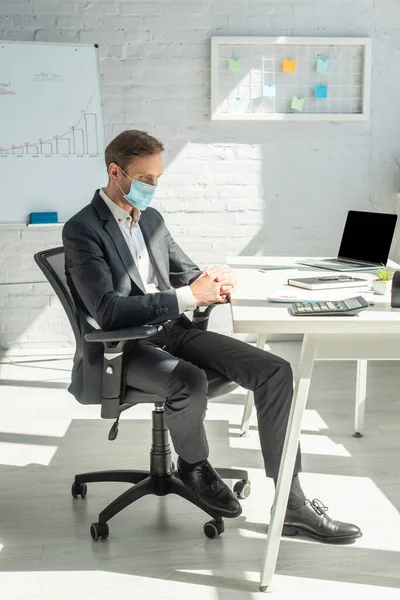 Longitud completa del hombre de negocios con las manos apretadas sentado en la silla de la oficina en el lugar de trabajo con el ordenador portátil y papelería en la mesa - foto de stock